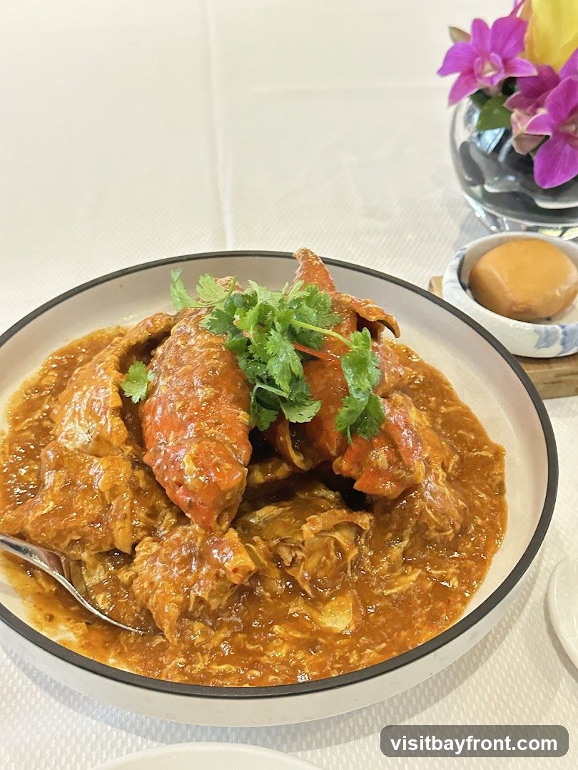 A close-up shot of a chili crab dish served in a white bowl with a black rim. The crab pieces are coated in a thick, orange-red sauce and garnished with fresh cilantro. A small side dish with a fried bun is visible to the right, along with a glass vase containing purple orchids and yellow flowers. The table is covered with a white tablecloth.