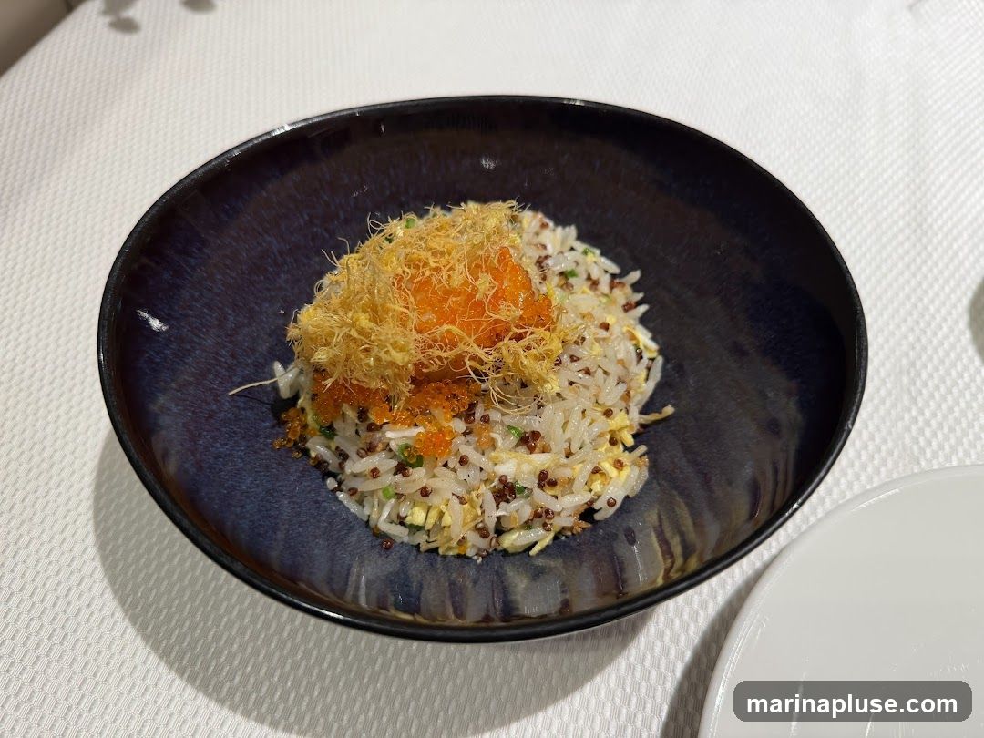 A close-up shot of a bowl of fried rice. The rice appears to be mixed with small grains, possibly quinoa, and flecks of egg and green onions. It is topped with a generous portion of shredded crispy fried egg floss and bright orange fish roe. The dish is served in a dark, iridescent blue bowl on a white tablecloth.