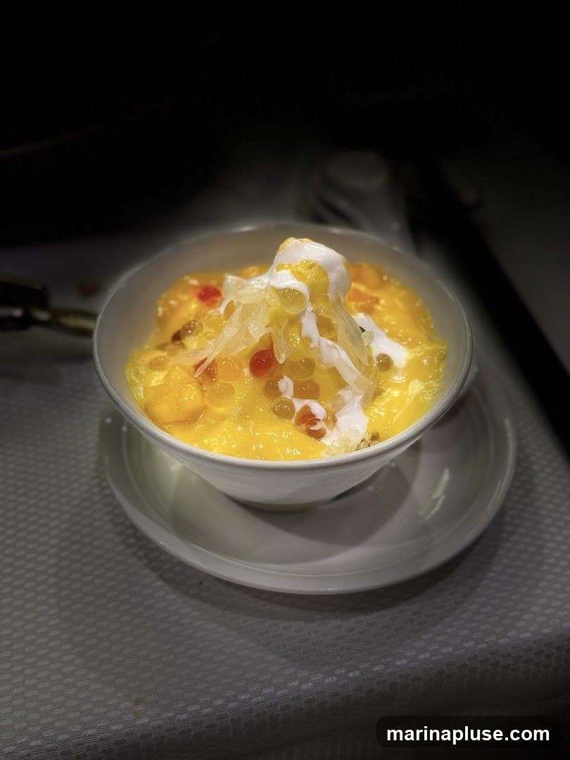 A close-up shot of a bowl of mango sago dessert, a popular Asian dessert. The dessert is served in a white bowl and consists of diced mangoes, sago pearls, and coconut milk, topped with a scoop of ice cream and shredded coconut. The dessert is presented on a white saucer and a textured tablecloth.