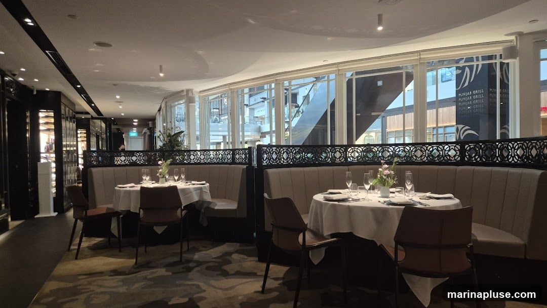 The image shows the interior of a restaurant with several tables set for dining. Booths with upholstered seating line the walls, separated by decorative screens. White tablecloths cover the tables, which are set with plates, cutlery, and glassware. In the background, through large windows, a shopping mall or atrium is visible, with escalators and other shops. The restaurant's name, "PUNJAB GRILL," is visible on signage outside the windows.