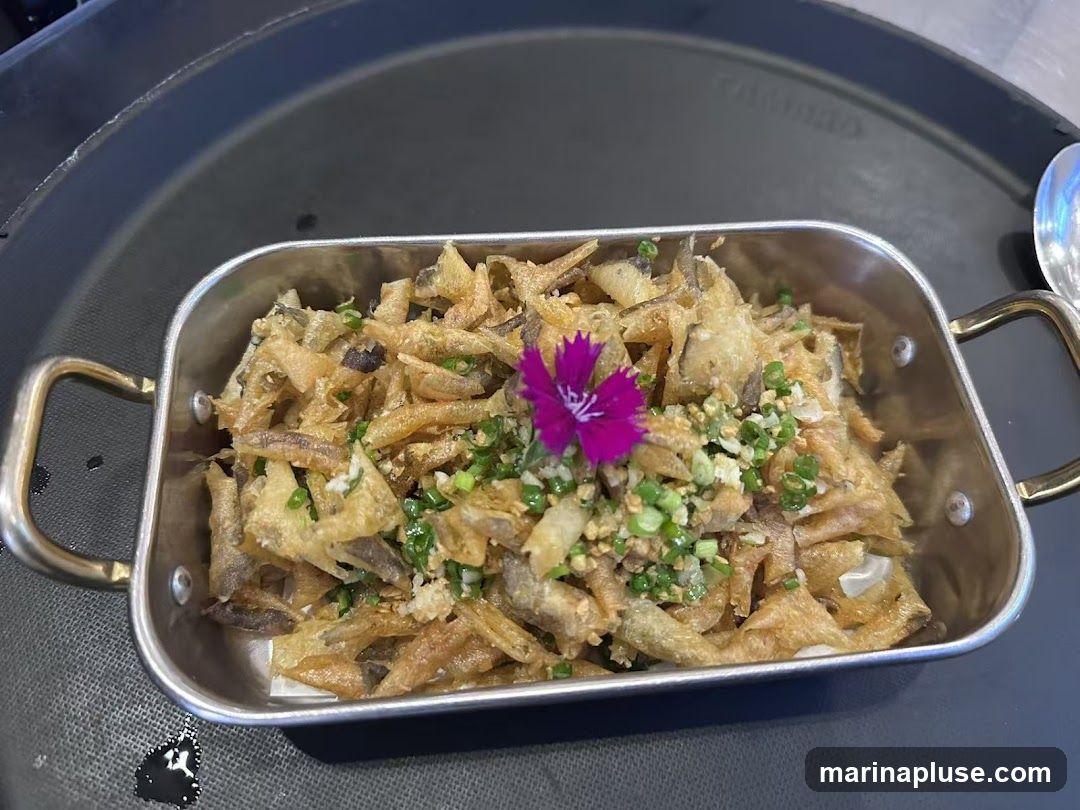 A close-up shot of a serving of crispy fried fish skin, garnished with chopped green onions and what appears to be minced garlic. A small, vibrant purple edible flower is placed on top as a garnish. The dish is presented in a rectangular metal container with handles, resting on a dark tray.