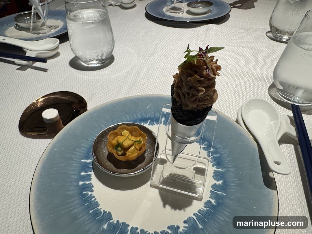 A close-up shot of a table setting with two small appetizers. In the foreground, a delicate pastry shell is filled with a creamy mixture and garnished with herbs. To its right, a black cone-shaped wafer holds a swirl of what appears to be a savory mousse, also topped with microgreens. The plates are white with a blue abstract pattern around the rim. Several glasses of water and chopsticks are also visible on the white tablecloth.