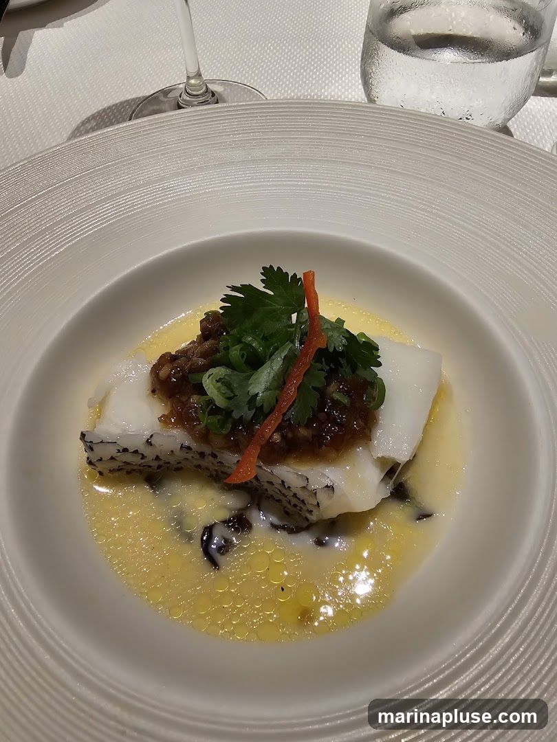 A close-up shot of a delicately prepared steamed fish dish served in a white, ribbed bowl. The fish, topped with a savory sauce, chopped scallions, and a sliver of red pepper, rests in a light, emulsified sauce. A glass of water and a wine glass are visible in the background, suggesting a fine dining setting.
