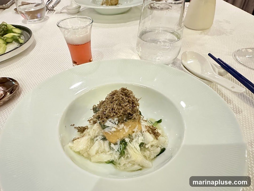A close-up view of a white plate with a delicate crab dish topped with finely shaved truffles. The dish appears to be served in a fine dining setting, with a small glass of pink liquid, a glass of water, chopsticks, and a spoon visible on the table. A side dish of asparagus is also present.