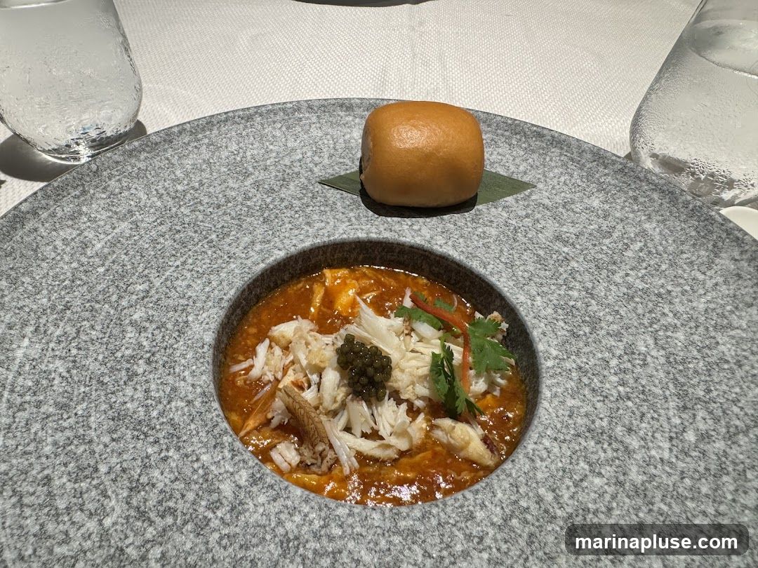 A close-up shot of a gourmet dish featuring shredded crab meat, caviar, and a rich orange sauce, served in a textured grey bowl. A small, golden-brown bun sits atop a green leaf on the side of the bowl. Two glasses of water are visible in the background, suggesting a dining setting.