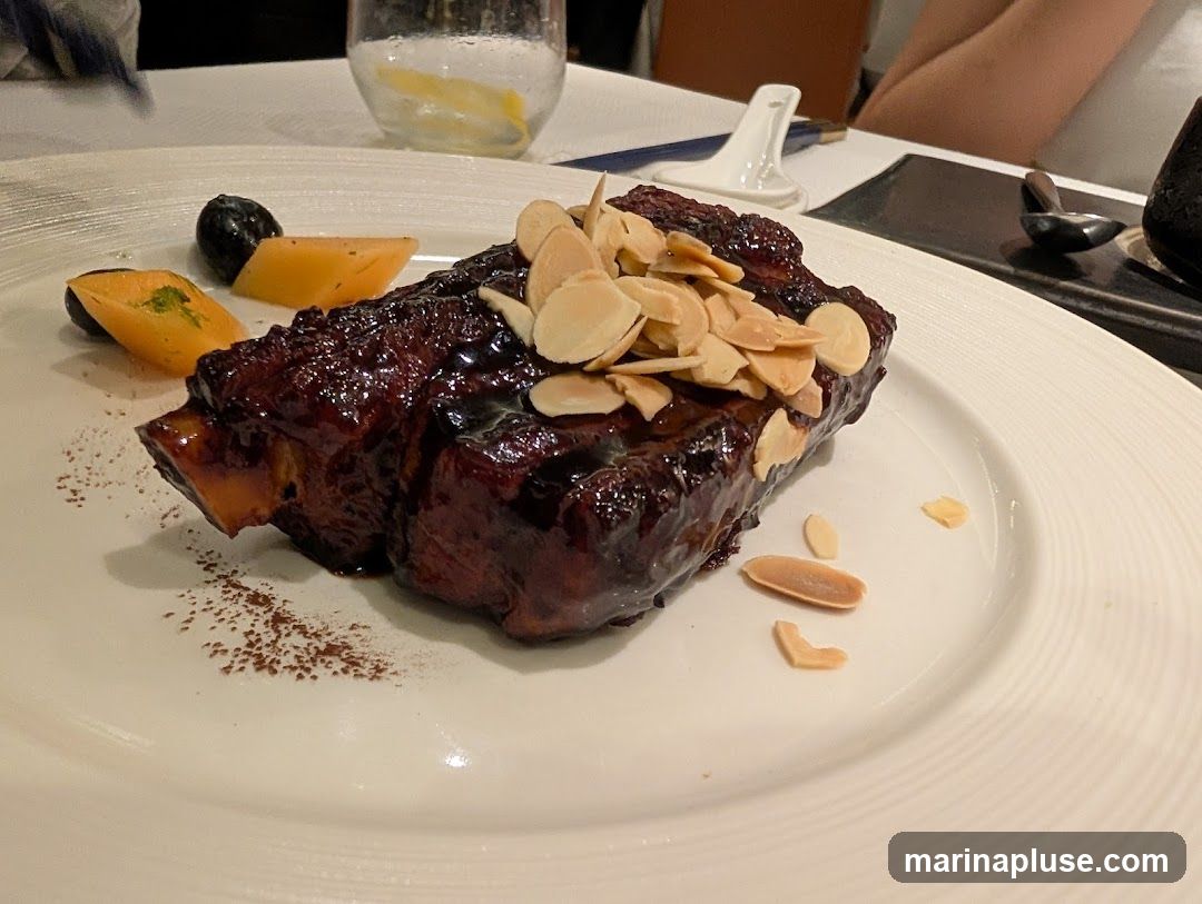 A close-up shot of a plate of braised short ribs, glazed with a dark sauce and topped with sliced almonds. The ribs are served with a few pieces of fruit, possibly mango, and a dark berry. A dusting of cocoa powder or spice is visible on the white plate. The setting appears to be a restaurant with a white tablecloth and a glass of water with lemon in the background.