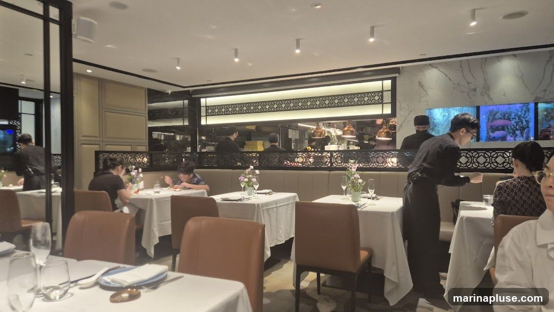 A view of a restaurant interior with tables set for dining. Several patrons are seated at tables, and staff members are visible, including one attending to a table and others in the kitchen area. The decor includes white tablecloths, brown chairs, and decorative screens. There are also aquariums with fish visible on the wall.