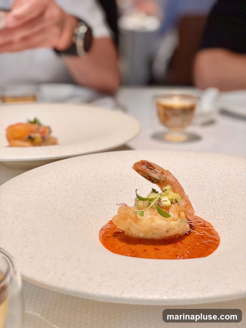 A close-up shot of a gourmet appetizer featuring a single fried shrimp served atop a mound of what appears to be a creamy filling, all resting in a vibrant red sauce. The dish is presented on a textured white plate. In the background, out of focus, are diners at a table, suggesting a restaurant setting.