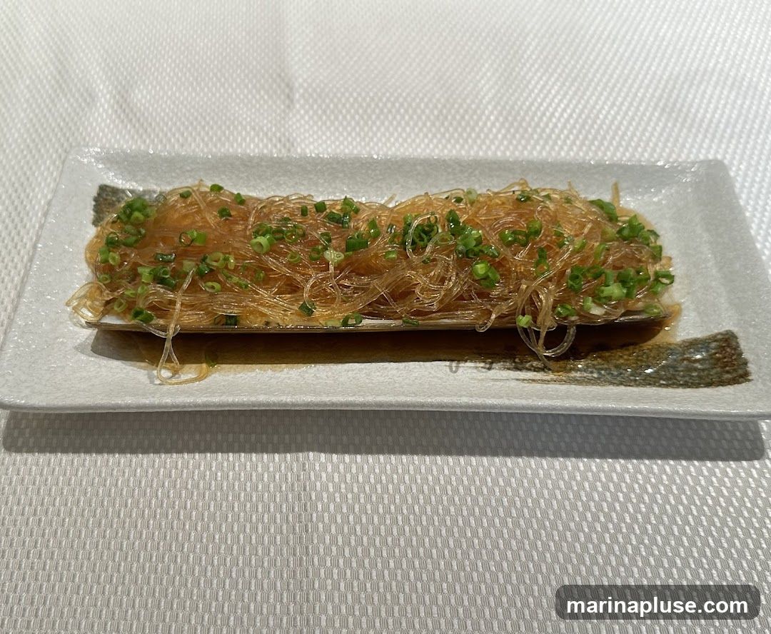 A rectangular white plate holds a serving of glass noodles, garnished with chopped green onions. The noodles are translucent and appear to be coated in a light sauce. The plate is set on a white tablecloth.