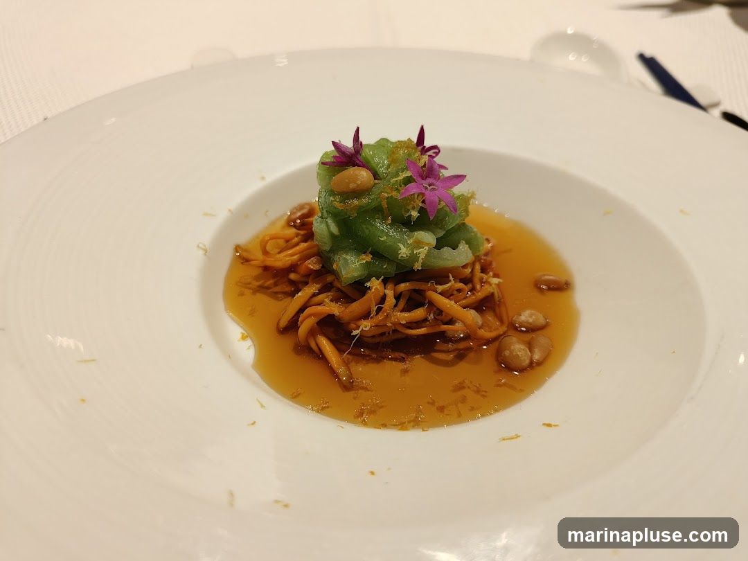 A close-up shot of a gourmet dish served in a white bowl. The dish features a mound of green vegetables topped with small purple flowers and garnished with citrus zest. It sits in a pool of amber-colored broth, surrounded by what appear to be cordyceps mushrooms and toasted nuts. The presentation is elegant and refined.