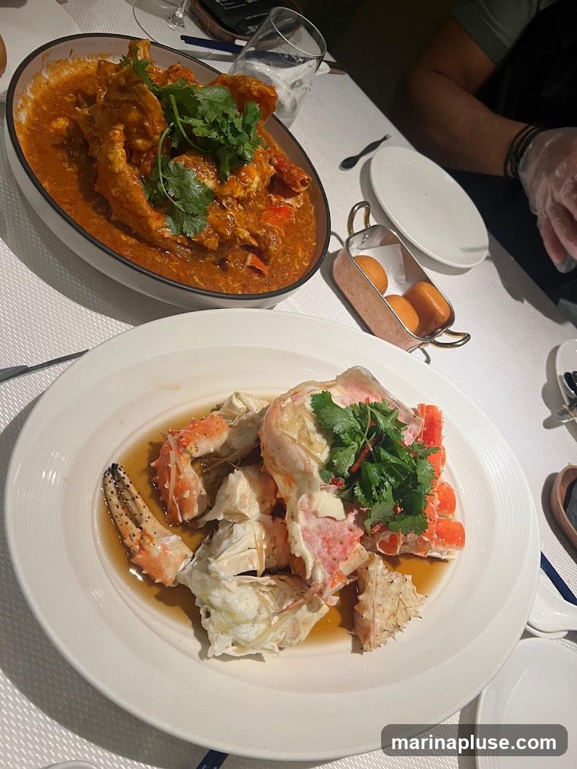 Two plates of crab dishes are presented on a white tablecloth. The dish in the foreground features cracked crab pieces in a light broth, garnished with cilantro. The dish in the background is a whole crab in a rich, orange sauce, also garnished with cilantro. A small copper dish with what appear to be fried buns or mantou is also visible, along with plates, cutlery, and glasses.