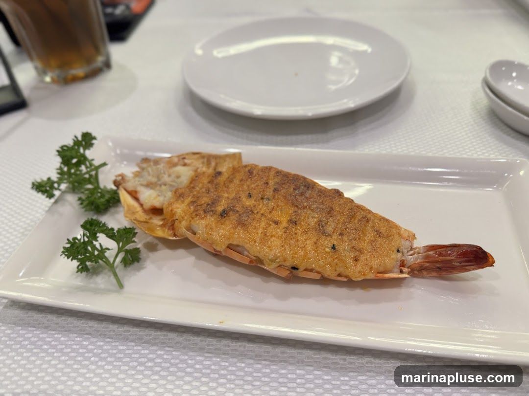 A close-up shot of a large baked shrimp served on a white rectangular plate. The shrimp is split lengthwise and topped with a golden-brown crust, possibly cheese or breadcrumbs. It is garnished with fresh parsley sprigs on the left side. In the background, a glass of dark liquid, a white plate, and a small white dish are visible on a white tablecloth.