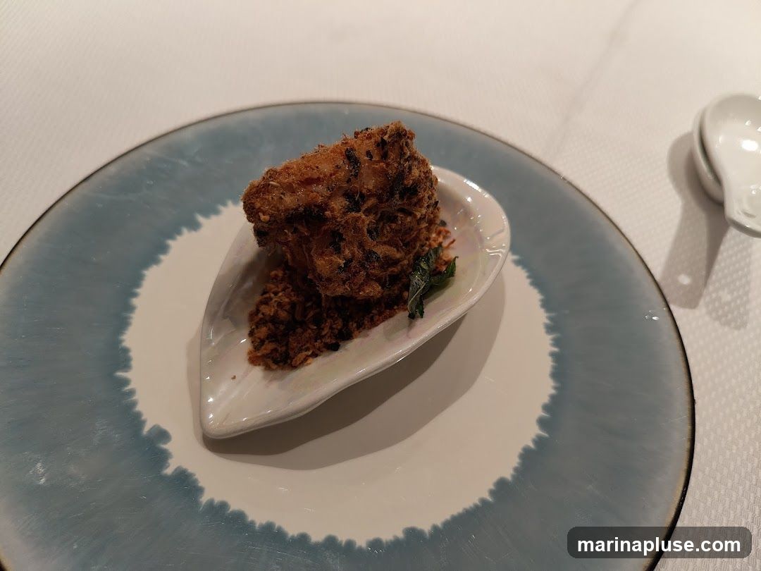 A close-up shot of a fried appetizer served on a small white dish, which is placed on a larger plate with a blue watercolor-like rim. The appetizer appears to be a piece of fried food coated in crispy, shredded material, served with a small garnish of green leaves. The plate is set on a white tablecloth. A white spoon is visible in the upper right corner.
