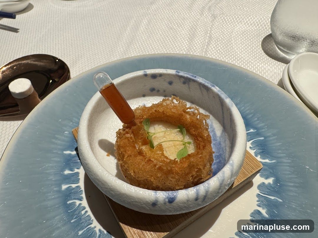 A close-up shot of a delicate appetizer served in a white bowl with blue accents, placed on a larger blue and white patterned plate. The appetizer features a crispy, golden-brown nest-like shell filled with a creamy white substance and garnished with a small green leaf. A small glass dropper filled with a reddish sauce is inserted into the nest. The table is set with a white tablecloth, a metallic spoon, and a glass of water.