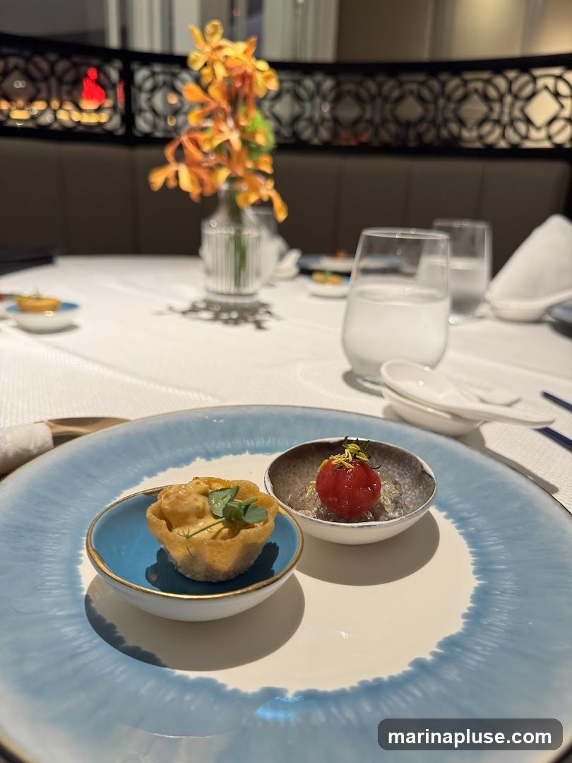 A close-up view of a table setting in a restaurant, featuring two small appetizers served on a large plate. The appetizer on the left is a small tart filled with a creamy mixture and garnished with microgreens. The appetizer on the right is a single cherry tomato topped with herbs, served in a small bowl. In the background, a vase with orange flowers adds a decorative touch. Two glasses of water and other tableware are visible, suggesting a formal dining experience. The table is covered with a white tablecloth.