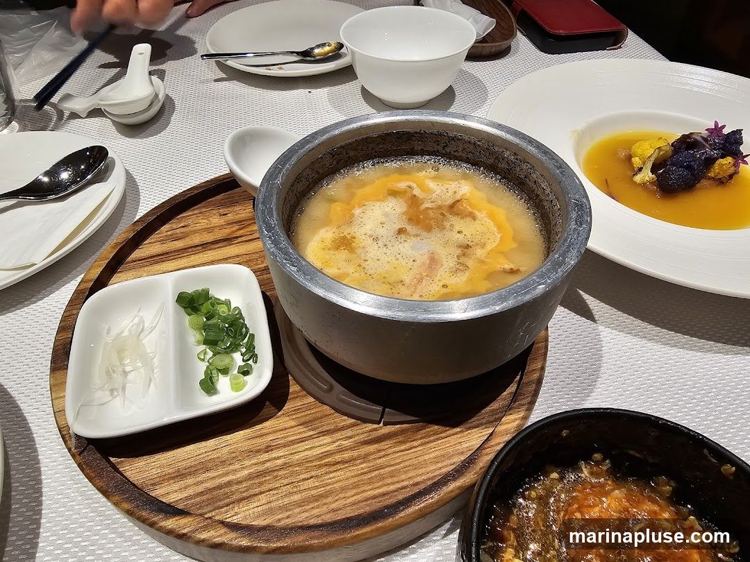 A close-up view of a table setting with various dishes. In the center, a metal pot contains a frothy, yellow soup. To its left, a small white dish is divided into two compartments, one holding thinly sliced white radish and the other chopped green onions. To the right of the soup pot, a white bowl holds a vibrant orange soup garnished with purple flowers and a piece of cauliflower. In the foreground, a dark bowl contains a saucy dish. The table is covered with a white textured tablecloth.