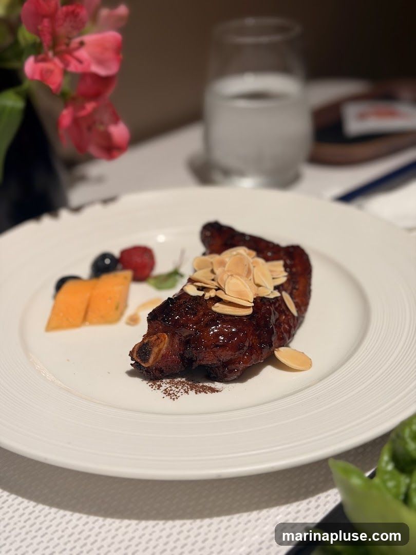 A close-up shot of a glazed rack of ribs, topped with sliced almonds, served on a white plate. The ribs are accompanied by a small garnish of cantaloupe slices, blueberries, and a raspberry. In the background, a glass of water and a vase of pink flowers are visible.