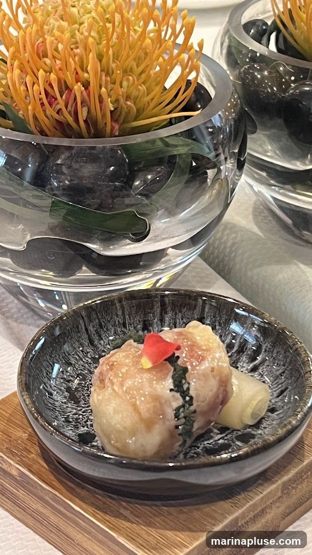 A close-up shot of a single appetizer served in a dark, textured bowl, resting on a wooden base. The appetizer appears to be a delicate pastry or dumpling, garnished with a single red and yellow petal. Behind it, a clear glass vase holds a vibrant orange flower and dark stones, with another similar vase visible in the background. The setting suggests a fine dining experience.