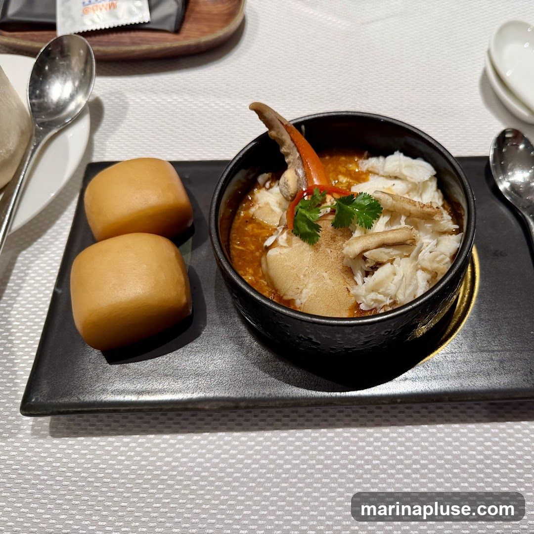 A close-up shot of a crab dish served in a black bowl, accompanied by two mantou buns on a black rectangular plate. The crab dish features a large crab claw and shredded crab meat in a rich, orange-colored sauce, garnished with cilantro and a sliver of red chili. The mantou buns are golden brown and appear soft. The setting suggests a restaurant meal, with silverware and a white tablecloth visible.
