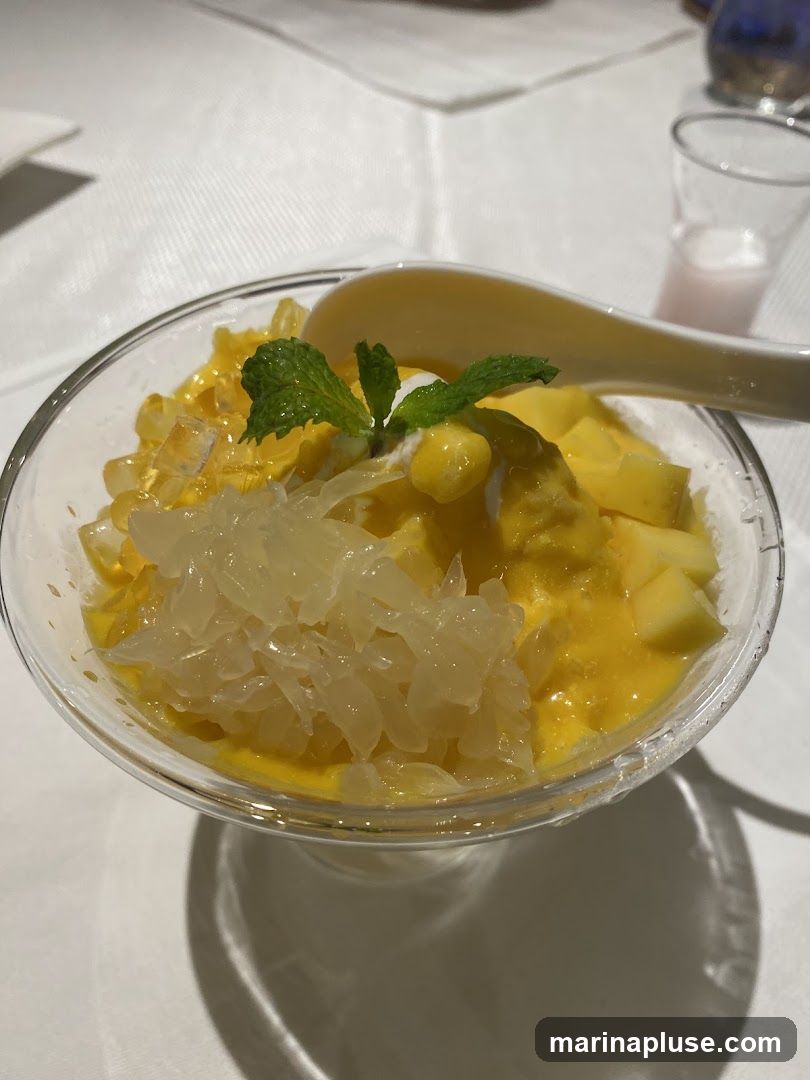 A close-up shot of a refreshing dessert in a clear glass bowl. The dessert features a base of creamy mango puree, topped with diced fresh mango, chunks of pomelo, and a sprig of mint. A white spoon rests on the edge of the bowl, ready to be used. A small glass with a pinkish liquid is visible in the background.