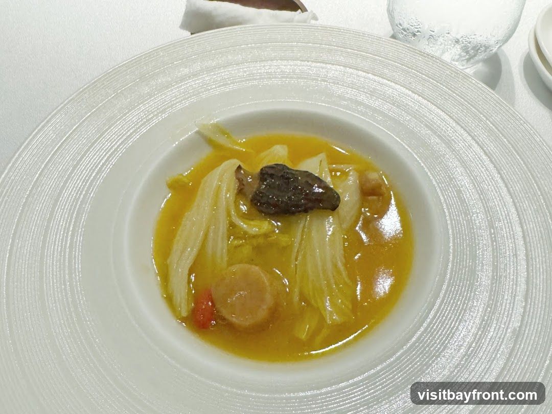 A close-up shot of a bowl of soup. The soup is a vibrant yellow broth filled with napa cabbage, a slice of mushroom, and a round piece of scallop. The bowl has a textured, concentric ring design. A glass of water is visible in the background.