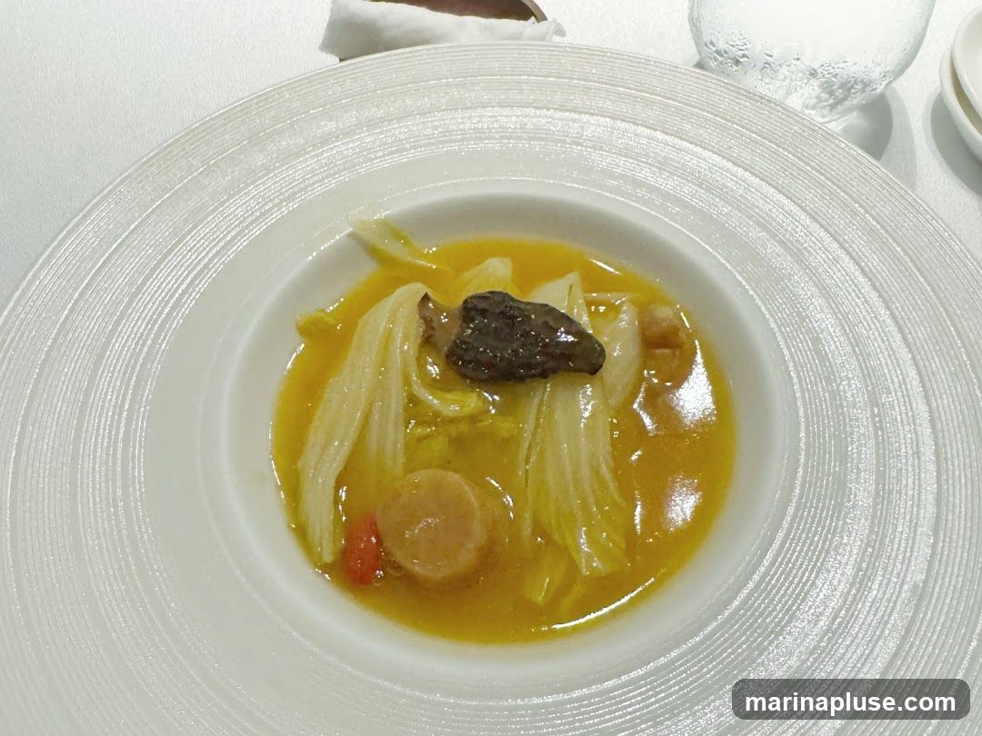 A close-up shot of a bowl of soup. The soup is a vibrant yellow broth filled with napa cabbage, a slice of mushroom, and a round piece of scallop. The bowl has a textured, concentric ring design. A glass of water is visible in the background.