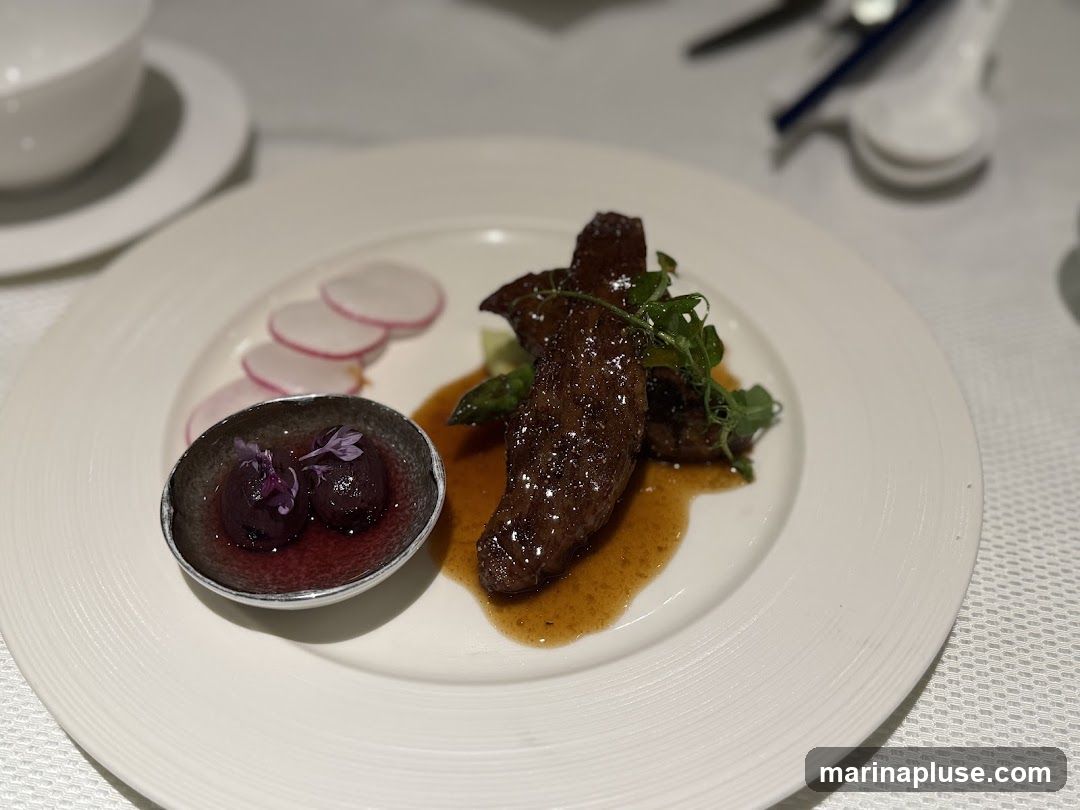 A close-up shot of a gourmet dish featuring several pieces of dark, glazed meat, possibly beef or lamb, served with a light brown sauce. Alongside the meat are a few sprigs of green garnish and a small dish containing two dark, round items, possibly berries or small fruits, in a red sauce, topped with delicate purple flowers. The dish is presented on a white plate with a textured rim, set on a white tablecloth. In the background, out of focus, are white bowls and utensils, suggesting a fine dining setting.
