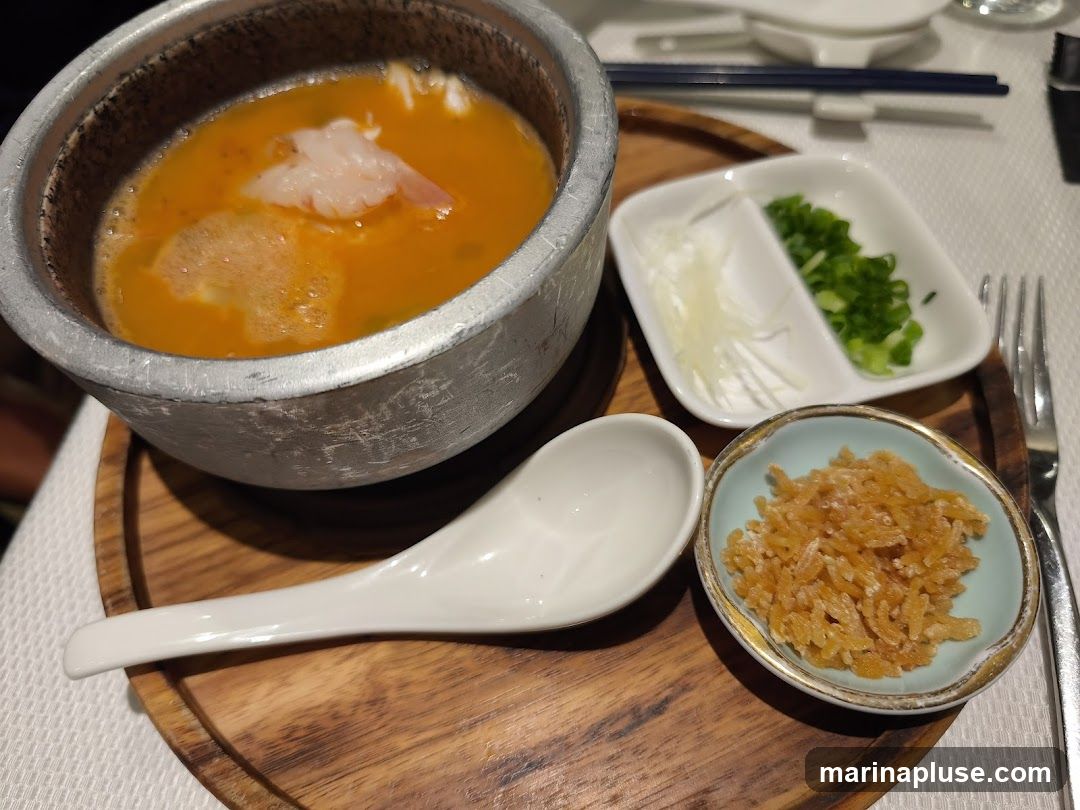 A close-up shot of a bowl of soup with a single shrimp and some foam. The soup is a vibrant orange color. To the right of the soup bowl are two small white dishes, one containing thinly sliced white ingredients and the other chopped green onions. In the foreground, a small blue dish holds a pile of small, crispy, golden-brown pieces. A white spoon rests on a wooden serving tray.