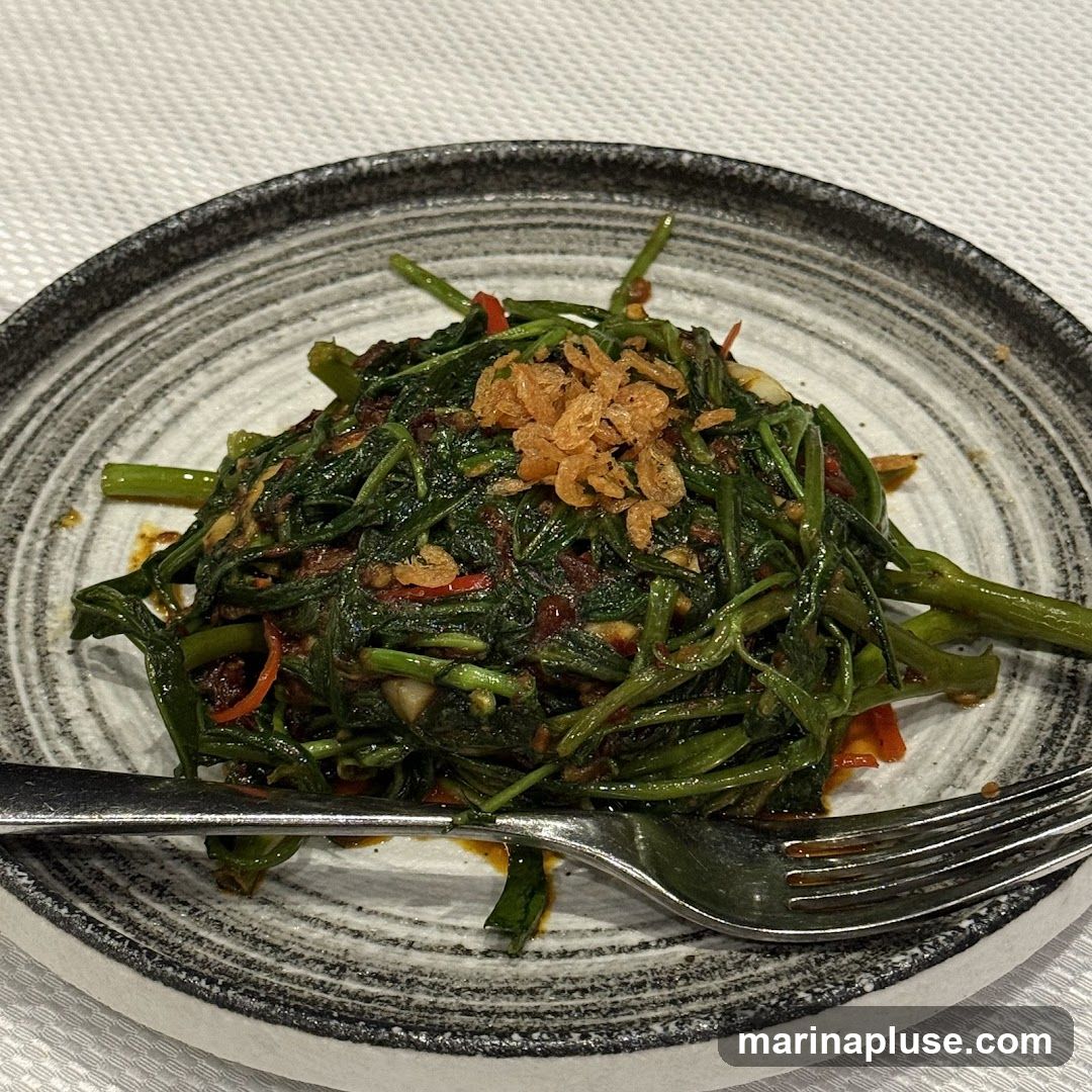 A close-up shot of a plate of stir-fried water spinach, also known as kangkong, seasoned with chili and garlic, and topped with crispy fried shallots. A fork rests on the side of the plate. The dish is served on a textured plate with a dark rim.