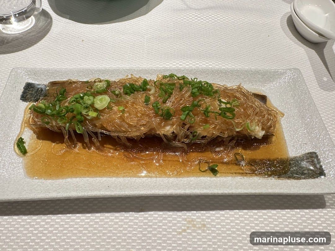 A rectangular white plate holds a dish of steamed fish topped with glass noodles and chopped green onions. The fish is served in a savory sauce. A small glass and a spoon are visible in the background.