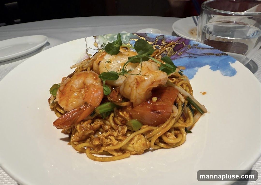 A plate of noodles topped with shrimp, scallops, and vegetables, garnished with microgreens. The plate has a decorative blue and gold rim. A glass of water is visible in the background.