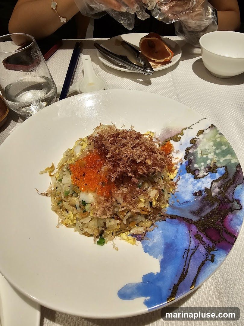 A plate of fried rice topped with fish flakes and orange roe is presented on a table. The plate has a decorative blue and purple abstract design on its rim. A glass of water, chopsticks, a spoon, and a nutcracker with a crab shell are also visible on the table. A person wearing plastic gloves is in the background.