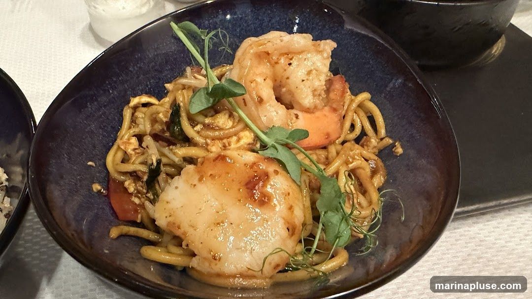 A close-up shot of a dark blue bowl filled with noodles, topped with a large shrimp and a seared scallop. The dish is garnished with microgreens.