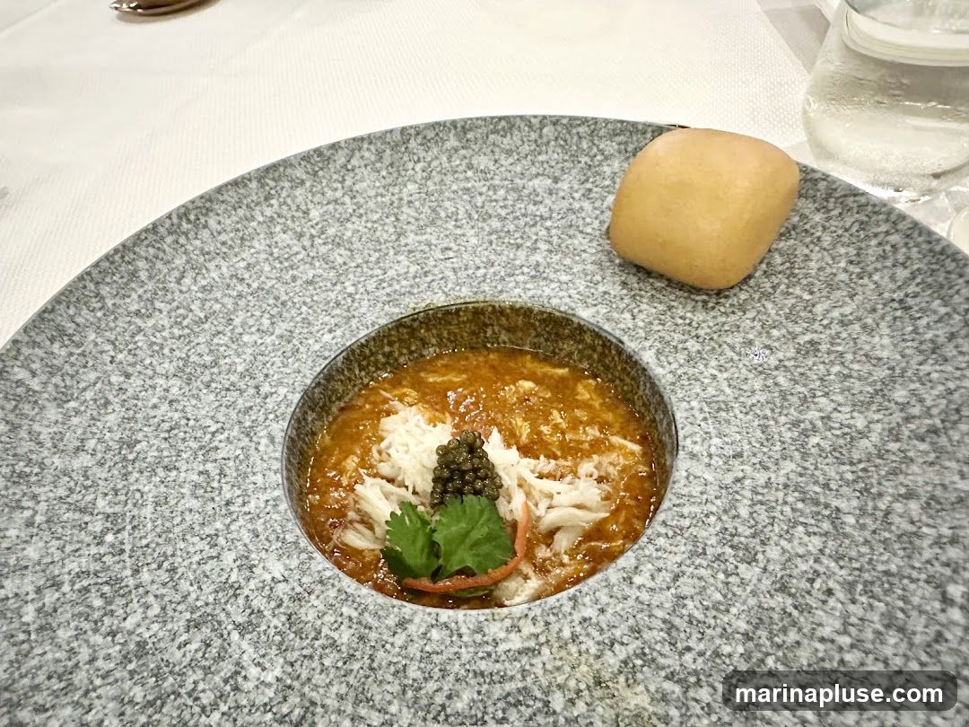 A bowl of crab bisque is served in a textured grey bowl, topped with shredded crab meat, a dollop of caviar, and a sprig of parsley. A small bread roll sits on the side of the large grey plate. A glass of water is visible in the background.