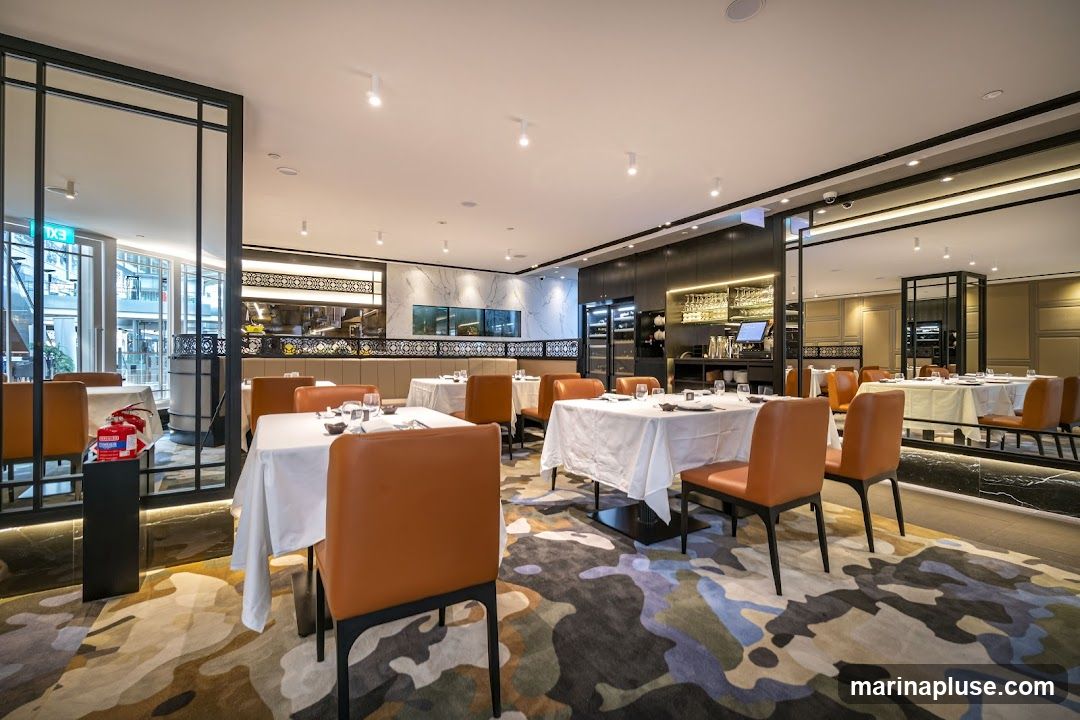 A well-lit, modern restaurant dining area with multiple tables set for service. The tables are covered with white tablecloths and set with place settings. Orange upholstered chairs surround the tables. The floor features a colorful, abstract patterned carpet. In the background, a bar area with wine displays and a kitchen pass-through are visible. Mirrors and decorative panels adorn the walls, creating a sense of spaciousness. An exit sign is visible through a glass partition on the left.