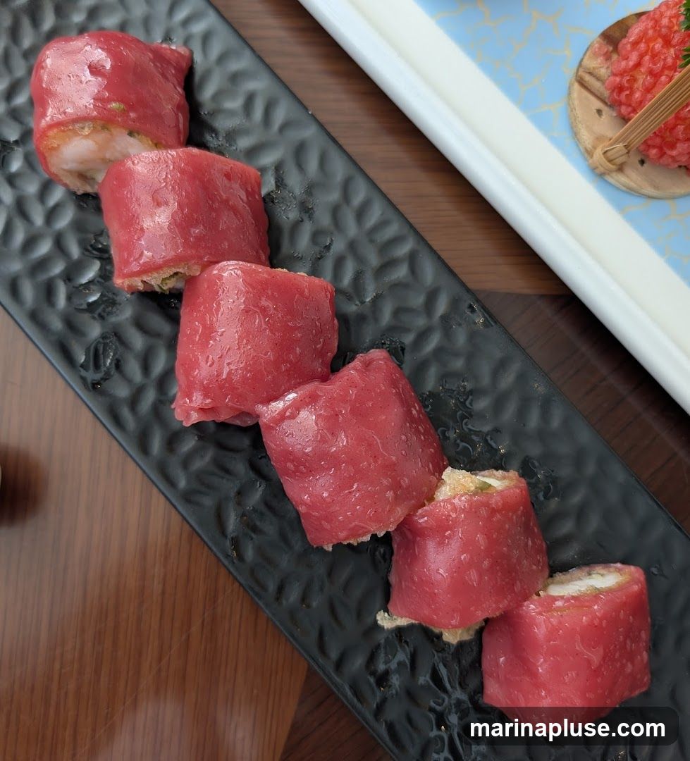 A close-up shot of six red-wrapped appetizers arranged diagonally on a black textured plate. The appetizers appear to be filled with a light-colored mixture, possibly seafood or vegetables. In the background, a white tray with a blue patterned rim holds a small dish with red, granular food and a bamboo skewer. The surface beneath the plate is a wooden table.