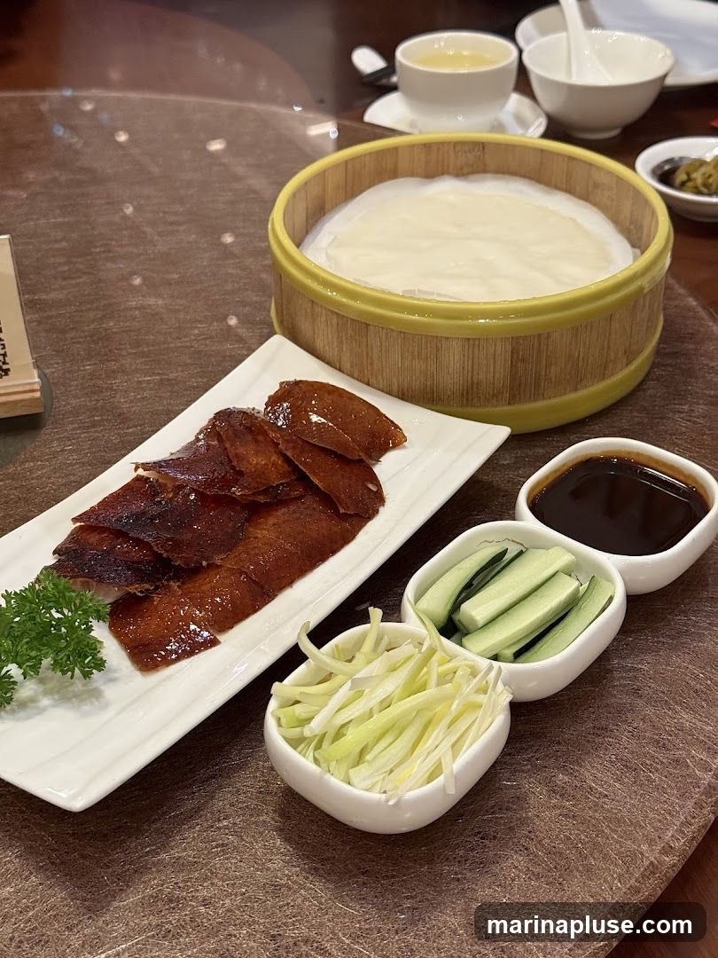 A close-up view of a Peking duck dish served with traditional accompaniments. The crispy duck skin is arranged on a white rectangular plate, garnished with a sprig of parsley. Alongside are small white bowls containing thinly sliced scallions, cucumber sticks, and a dark dipping sauce. A bamboo steamer basket filled with thin pancakes sits in the background, along with a bowl of soup and other condiments.