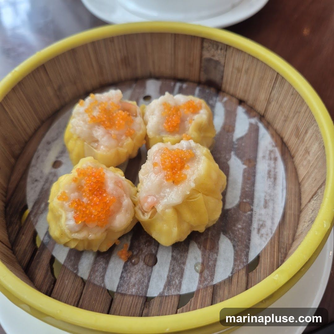 A close-up shot of four shrimp dumplings, also known as siu mai, served in a bamboo steamer basket. The dumplings are topped with bright orange roe.