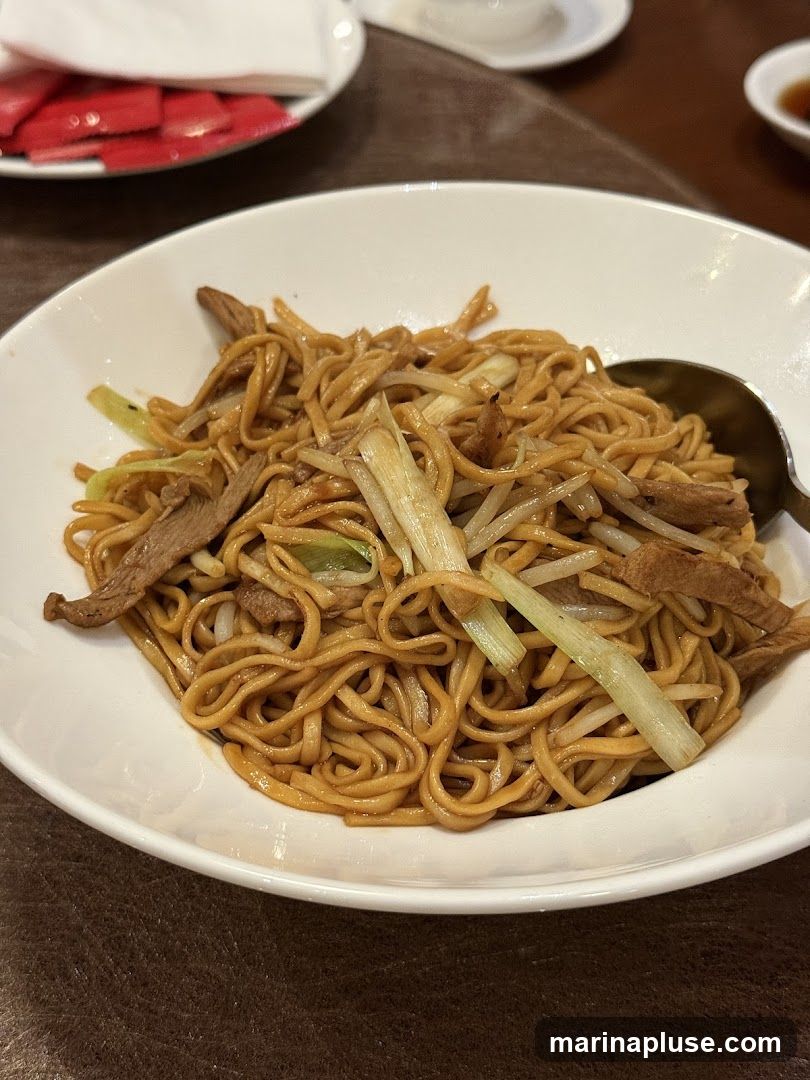 A close-up shot of a white bowl filled with lo mein noodles, thinly sliced meat, and green onions. A spoon rests on the right side of the bowl. In the background, out of focus, are red packets and white plates.