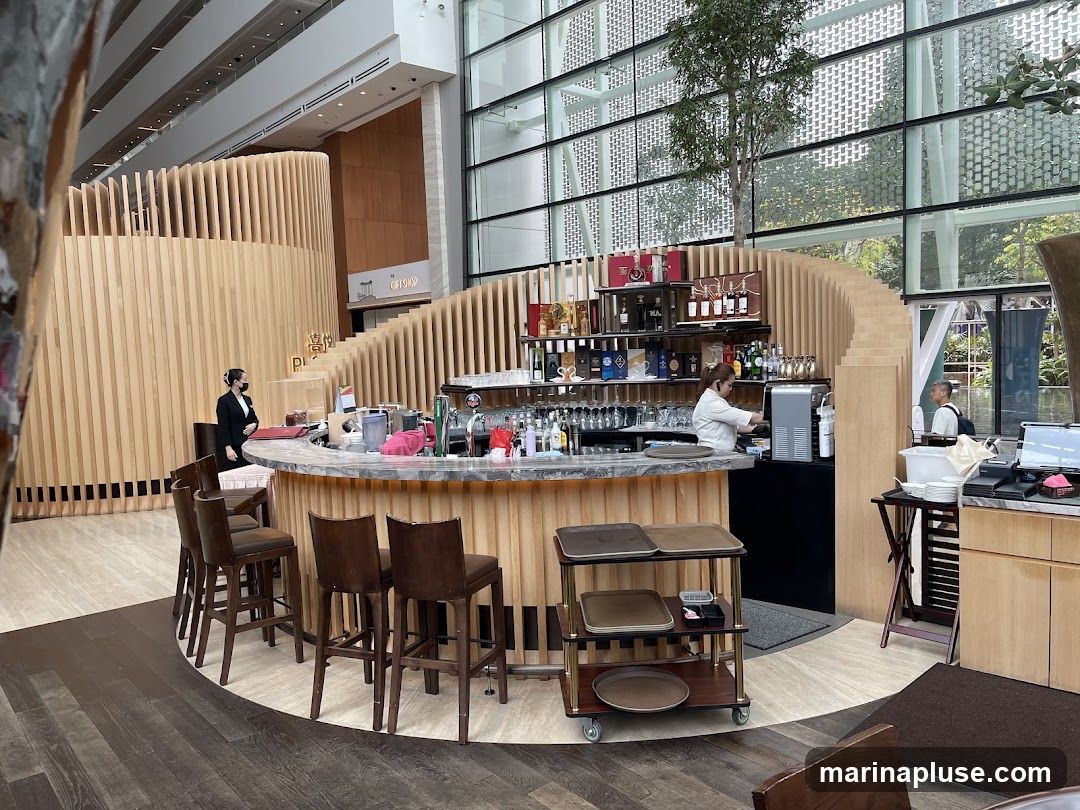 A modern bar area with a curved counter and wooden slatted walls. Two staff members are visible, one behind the counter preparing drinks and another standing near the entrance. Bar stools are arranged around the counter. Shelves behind the bar are stocked with bottles. A spiral staircase is visible in the background.