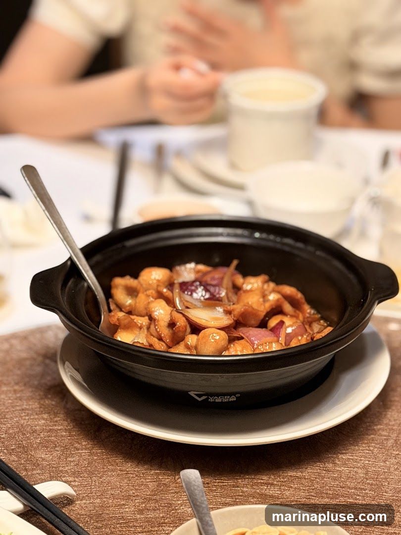A close-up shot of a dish featuring pieces of chicken and red onion in a savory sauce, served in a black ceramic pot with a spoon. The pot is placed on a white saucer on a textured brown placemat. In the background, out of focus, are diners at a table with white tableware and containers.