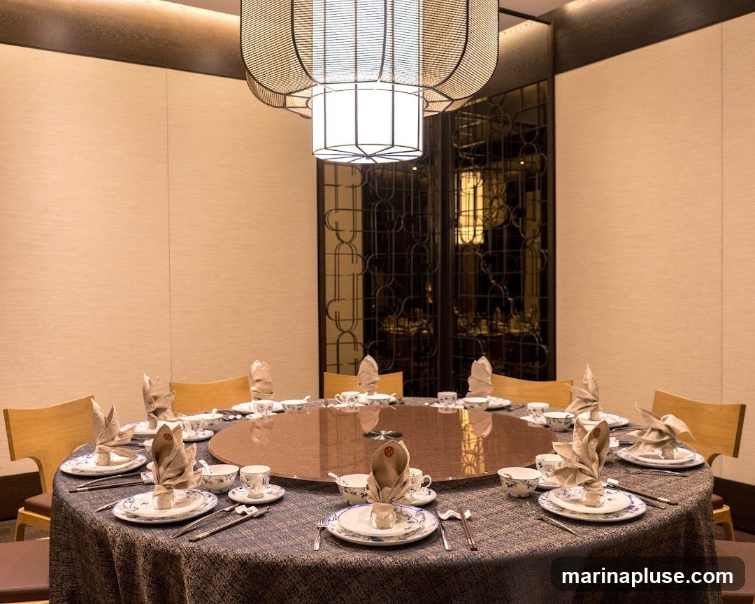 A round dining table is set for a formal meal in a well-lit room. The table is covered with a patterned tablecloth and features place settings for multiple guests, each with plates, cutlery, and folded napkins. A large, decorative pendant light hangs from the ceiling above the table. The background shows a wall with subtle texture and a decorative screen.