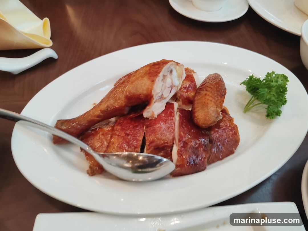 A platter of sliced roasted chicken is presented on a white oval plate. A spoon rests on the plate, and a sprig of parsley garnishes the dish. The chicken has crispy, golden-brown skin and appears to be juicy. The setting suggests a restaurant meal.