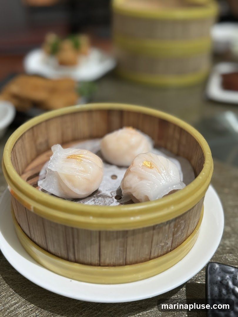 A close-up shot of three steamed dumplings in a bamboo steamer basket. The dumplings are translucent with visible shrimp filling and topped with a small piece of gold leaf. The steamer basket is placed on a white plate, and other blurred dishes are visible in the background.