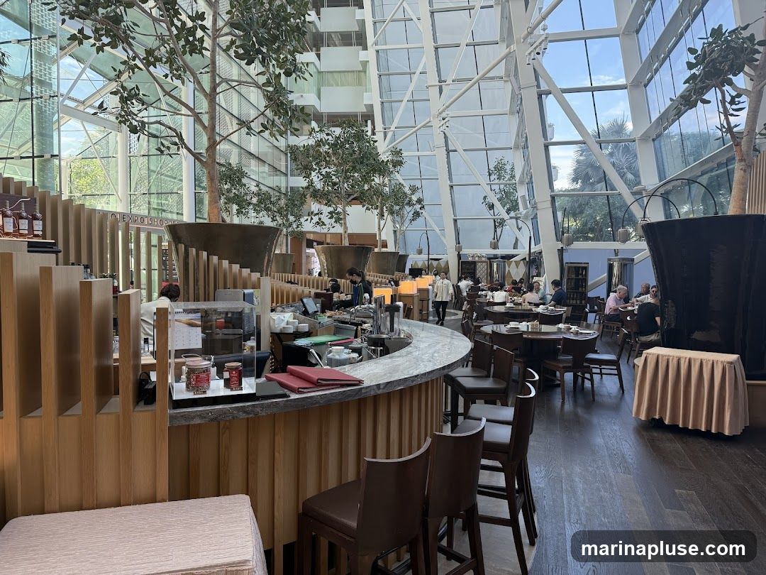 A spacious restaurant interior with a high, glass-paneled atrium ceiling. Large trees are placed throughout the dining area, which features round tables with patrons seated. A bar area is visible in the foreground with stools.