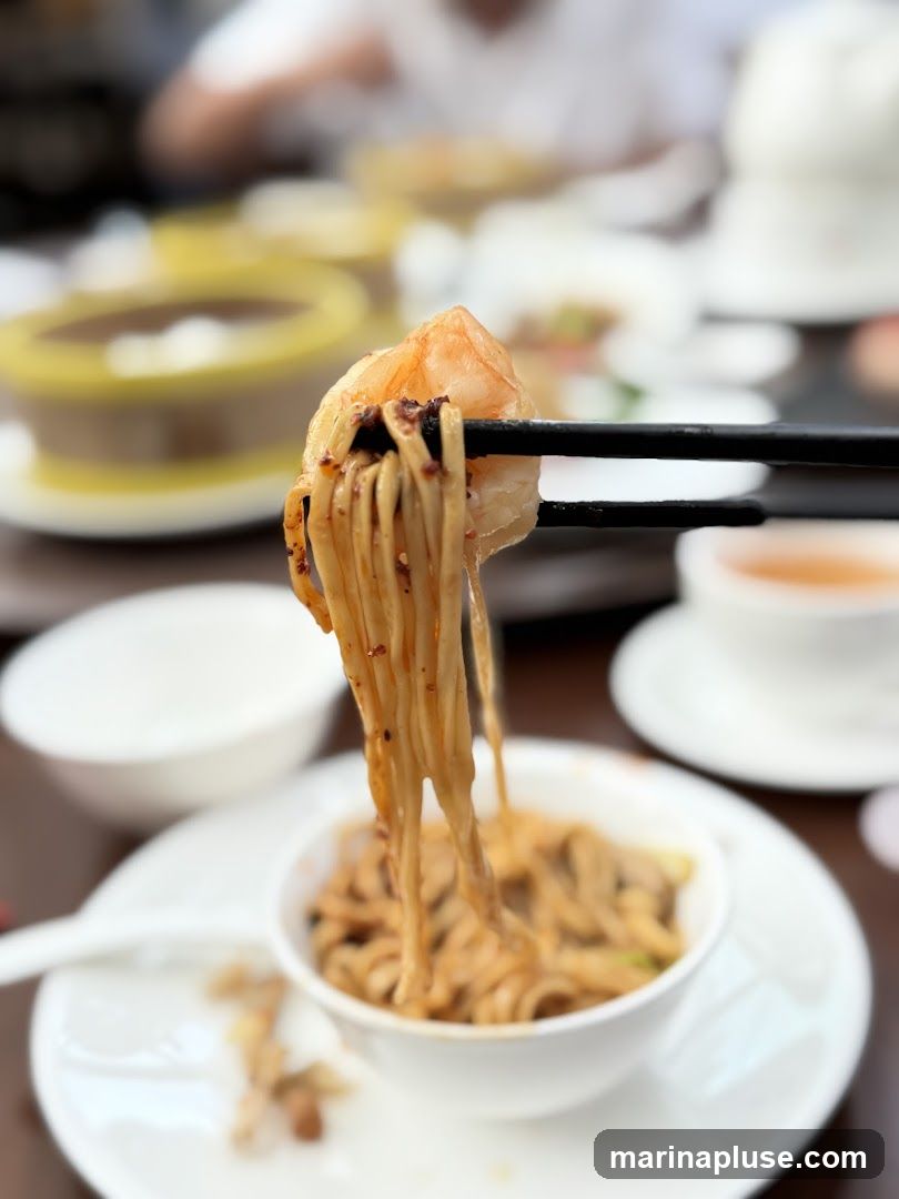 A close-up shot shows chopsticks lifting a portion of noodles with a shrimp from a bowl. The noodles appear to be coated in a savory sauce. In the background, other dishes and diners are out of focus.