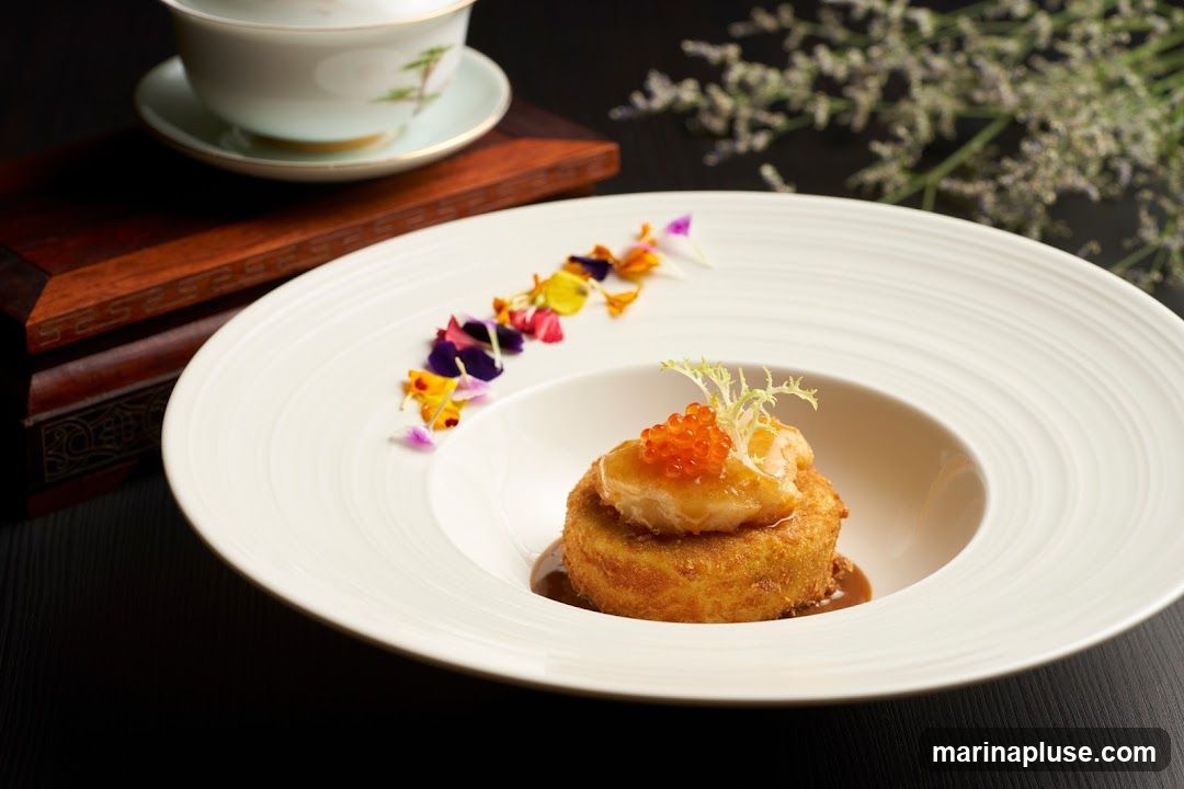A close-up shot of a beautifully presented dish. The main element is a golden-brown, crispy-coated round item, topped with a piece of seafood, roe, and a sprig of greens. It sits in a white, wide-rimmed bowl with a dark sauce. The dish is garnished with colorful edible flower petals arranged in a curve. In the background, a traditional tea cup and saucer are visible on a wooden stand, with delicate white flowers adding a soft touch to the dark backdrop.