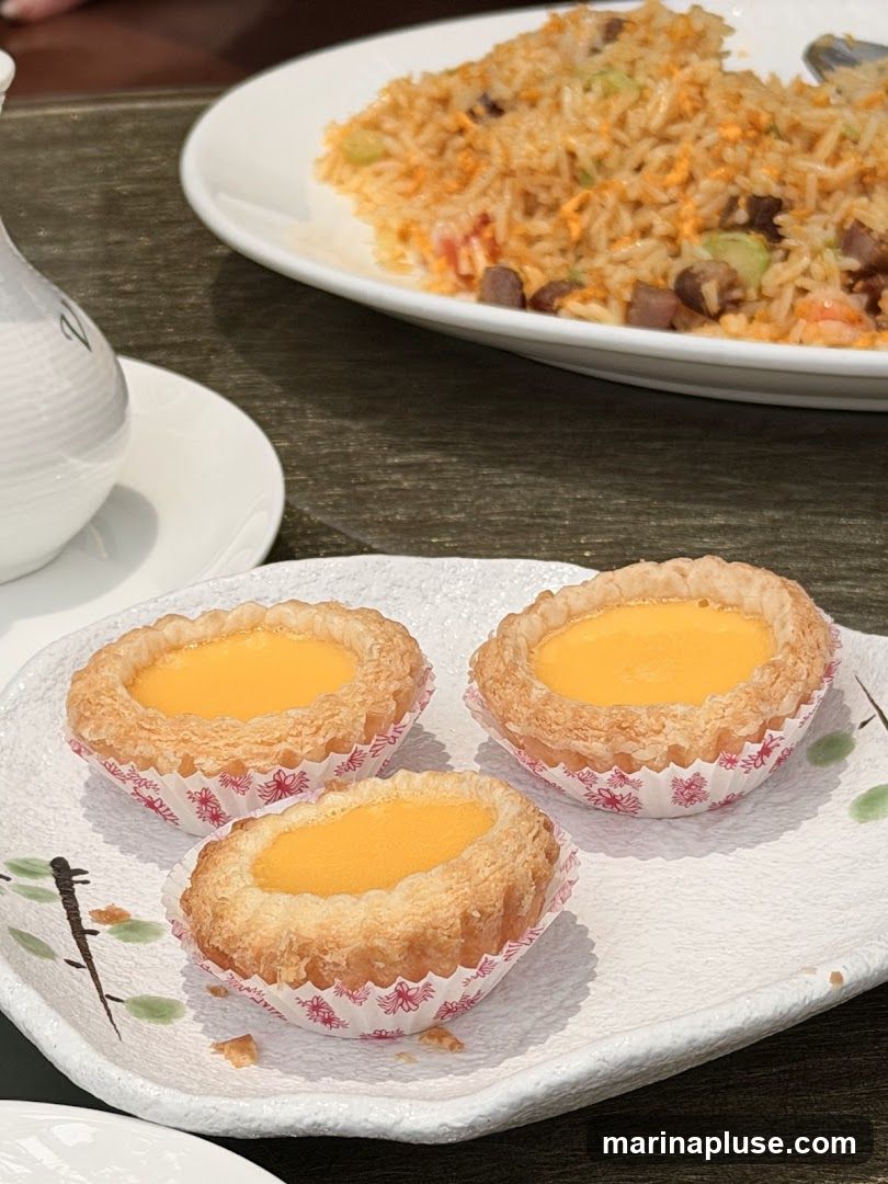 Three golden egg tarts sit on a decorative white plate with a subtle floral design. In the background, a plate of fried rice is visible, featuring visible pieces of meat and vegetables. A white teapot or creamer is also partially visible on the left.