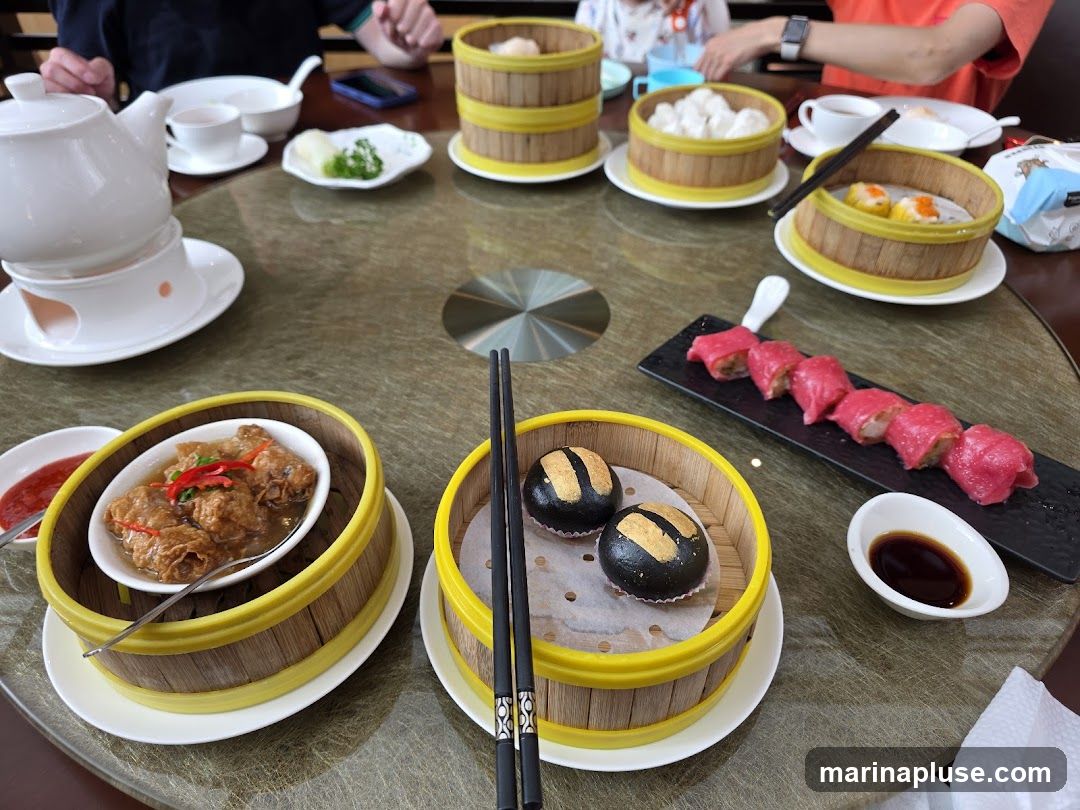A round table laden with various dim sum dishes, including steamed buns, dumplings, and a dish of braised tofu skin. A teapot and tea cups are also visible, suggesting a tea ceremony. The meal is being enjoyed by several people, whose hands and arms are visible around the table.