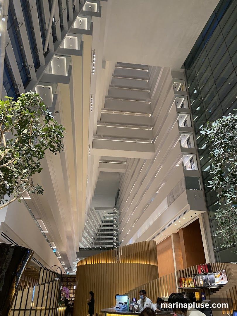 A wide-angle shot looking up into a vast, modern atrium with multiple levels of balconies and a glass exterior wall. Lush green trees are visible on the left and right sides, adding a touch of nature to the architectural space. In the foreground, a circular wooden bar area is illuminated, with several people, likely staff and patrons, interacting. The overall impression is one of a spacious and contemporary hotel lobby or a large commercial building interior.